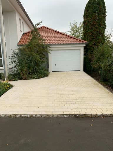 Helles Pflaster vor einer Garage mit begrünten Rändern und einem roten Dach.