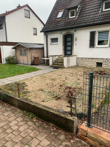 Einfamilienhaus mit Garten, Terrasse und kleiner Garage, umgeben von einem Zaun.