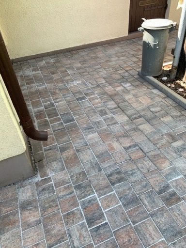 Gepflasterter Boden mit grauen und braunen Steinen, daneben ein Mülleimer.