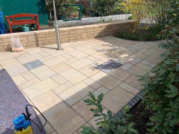 Gestreute Fliesenfläche mit Pflanzen und einer Bank im Hintergrund.