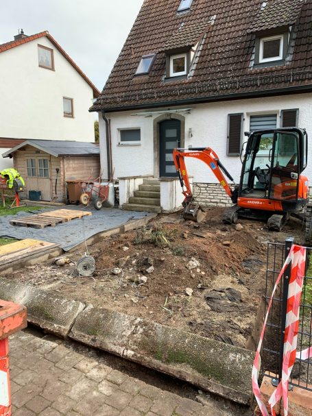 Baustelle mit Bagger, unebenem Gelände und Haus im Hintergrund. Baustellenabsperrung sichtbar.