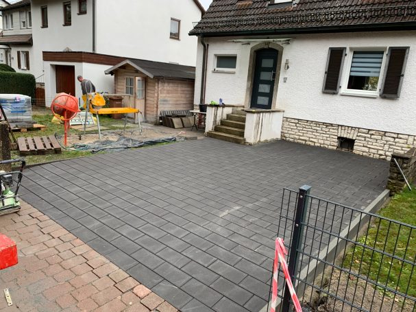 Pflasterfläche vor einem Haus mit Garten und Baugeräten im Hintergrund.