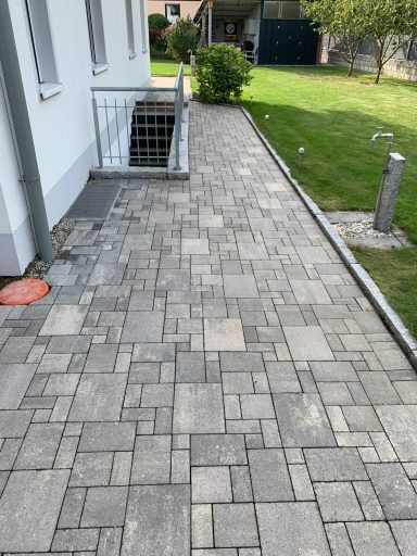 Schlichter Gehweg aus Steinen neben einem Haus, umgeben von Rasen und Bäumen.